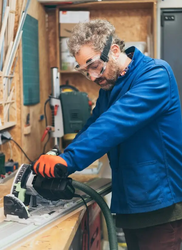 Un homme travaille dans un atelier menuiserie, un homme utilise une scie plongeante, il porte des EPI